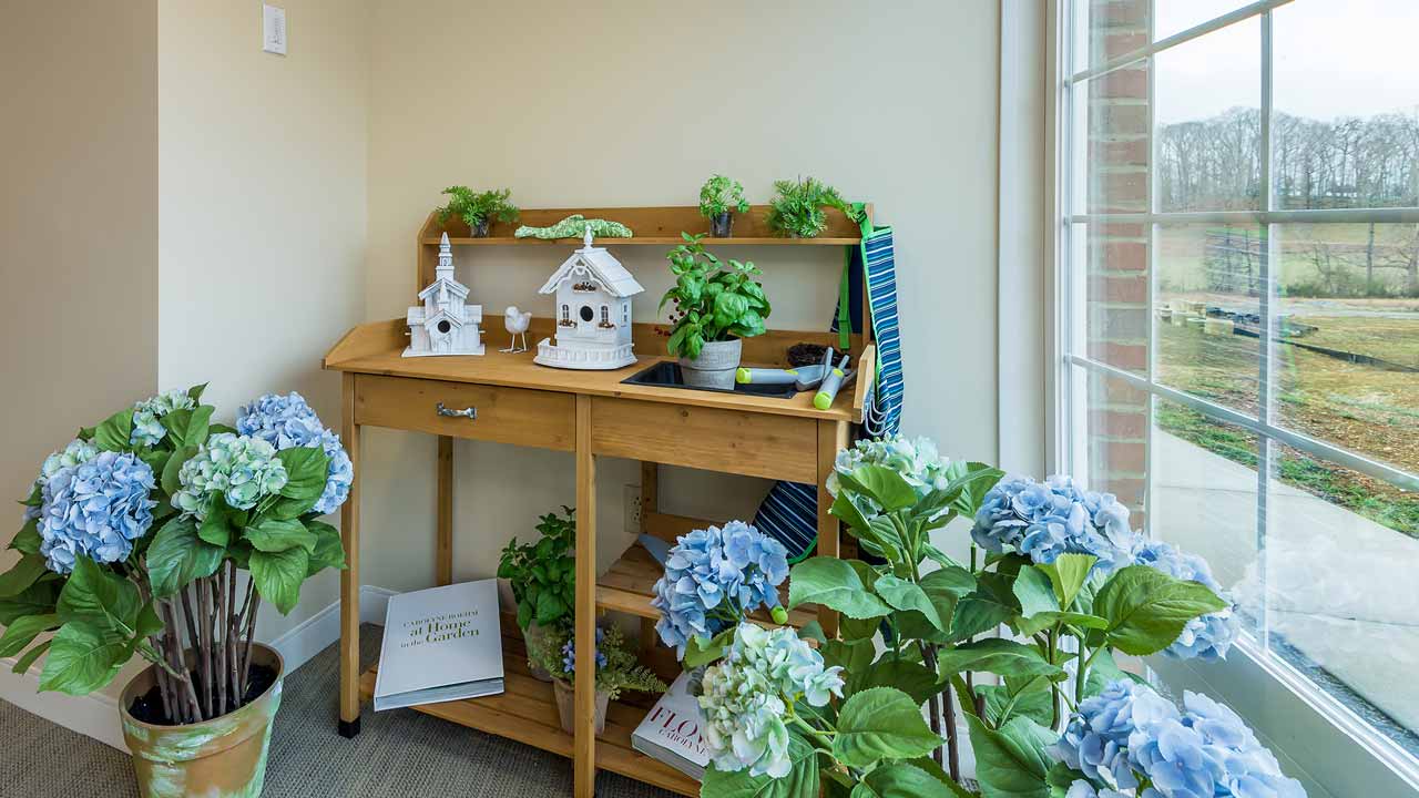 A wooden potting bench with potted plants, birdhouses, and gardening tools sits by a large window. Blue hydrangeas in pots decorate the area, and an open gardening book lies on the floor.