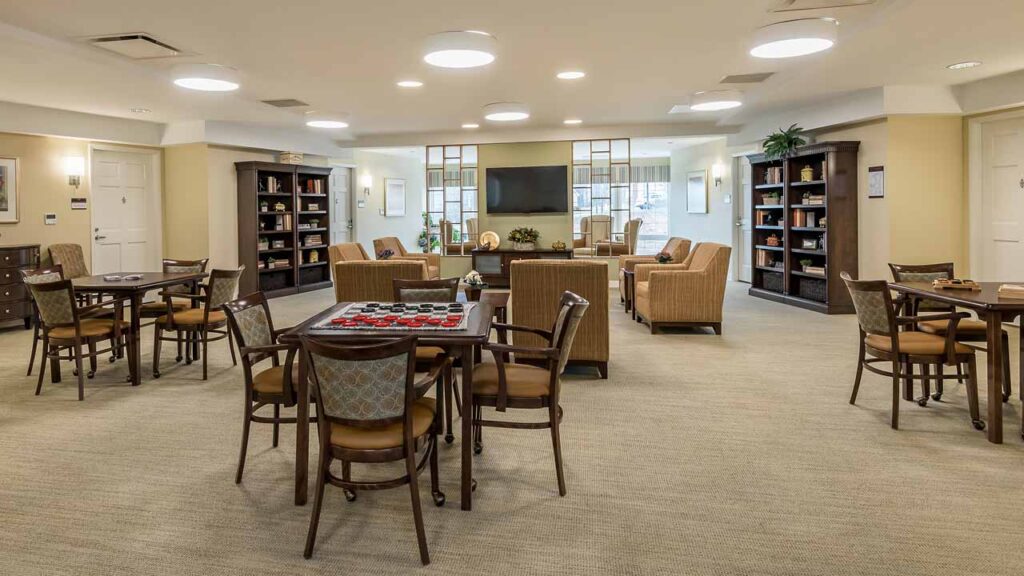 Spacious, well-lit community room with tables and chairs, a checkers game set up, lounge seating area, bookshelves, plants, and a large TV on the back wall. The atmosphere is cozy and inviting.