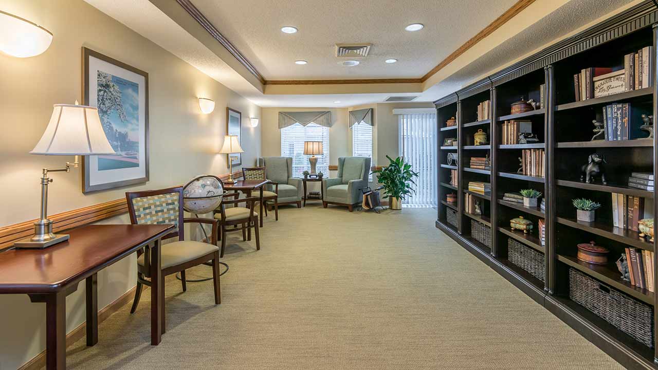 A cozy library room with comfortable chairs near windows, wooden tables and chairs, framed artwork on the wall, and a large bookshelf filled with books and decorative items. Warm lighting creates an inviting atmosphere.