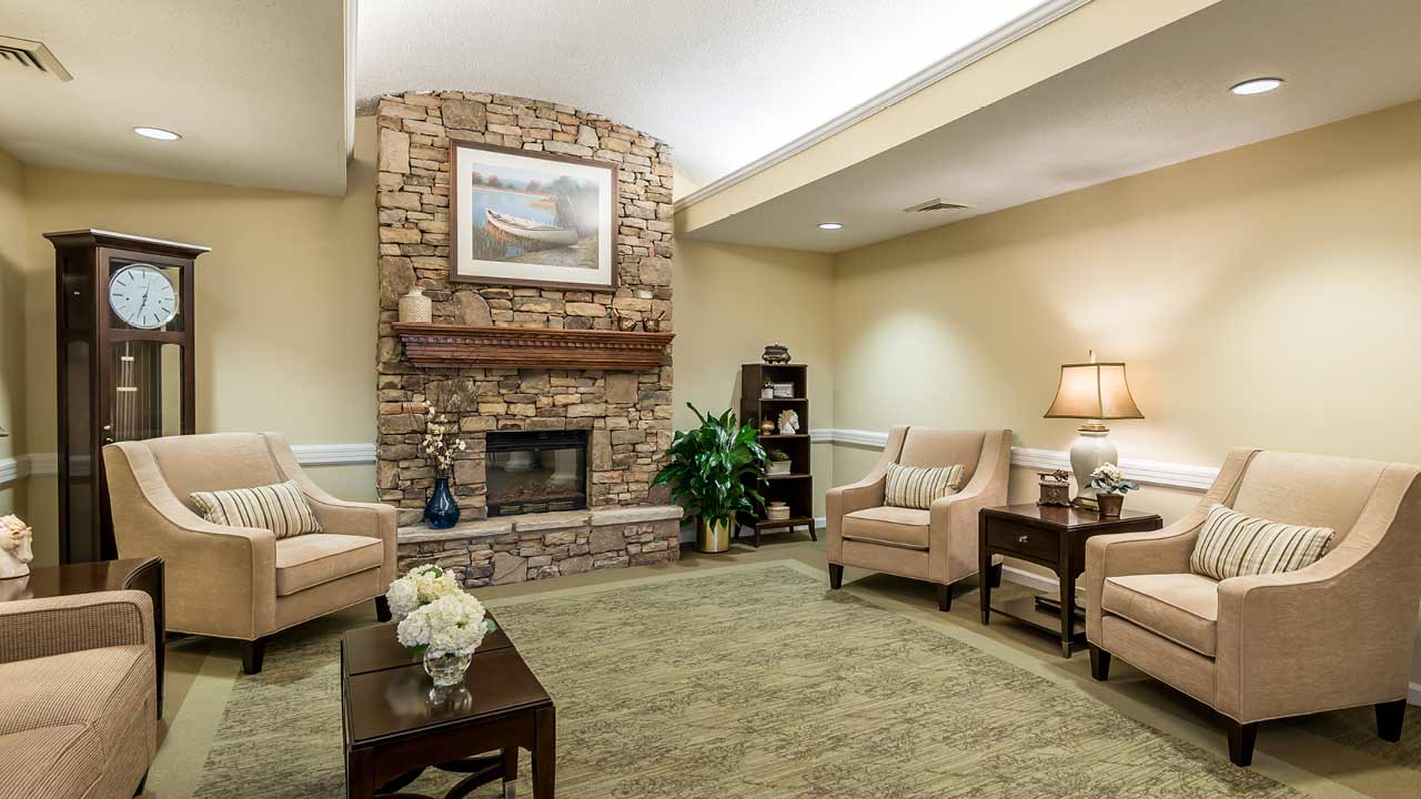 A cozy living room with beige armchairs, a stone fireplace, a wooden mantel, a grandfather clock, side tables with lamps, and decorative plants, all under soft ceiling lights.
