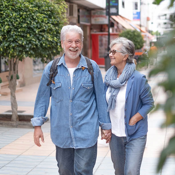An older couple walks hand in hand down a sunny city street, smiling and wearing casual blue clothing. They appear happy and relaxed, enjoying each other's company. Trees and shops line the background.