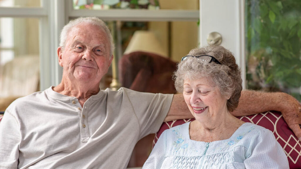 An elderly man and woman sit together on a couch. The man is smiling at the camera, while the woman, wearing glasses on her head, looks down and smiles. They appear relaxed and content in a cozy indoor setting.