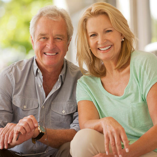 An older man and woman sit closely together indoors, both smiling warmly at the camera. The man wears a gray shirt and the woman wears a light green top. Sunlight filters in through the window behind them.