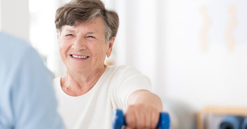 An older woman with short gray hair smiles while lifting a blue dumbbell indoors, wearing a white shirt. The background is softly blurred, creating a bright and positive atmosphere.