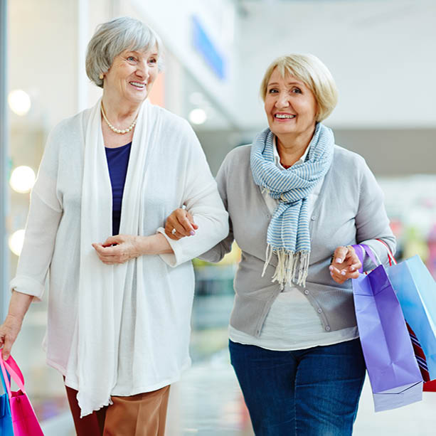 Two older women smiling and walking arm in arm in a mall, each holding colorful shopping bags, appearing happy and enjoying their shopping trip together.