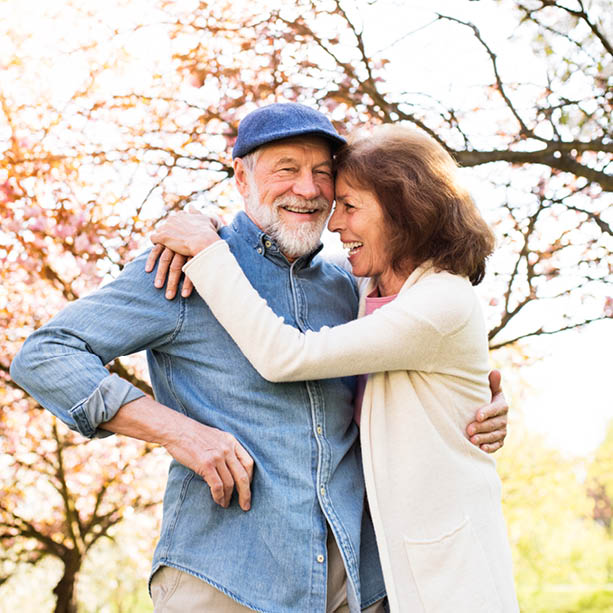 An older couple embraces and laughs together outdoors under blossoming trees, enjoying a sunny day. The man wears a blue cap and denim shirt; the woman wears a light cardigan.