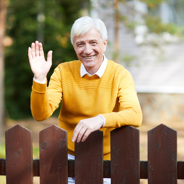 An older man with white hair, wearing a yellow sweater, smiles and waves while standing behind a brown wooden fence outdoors.