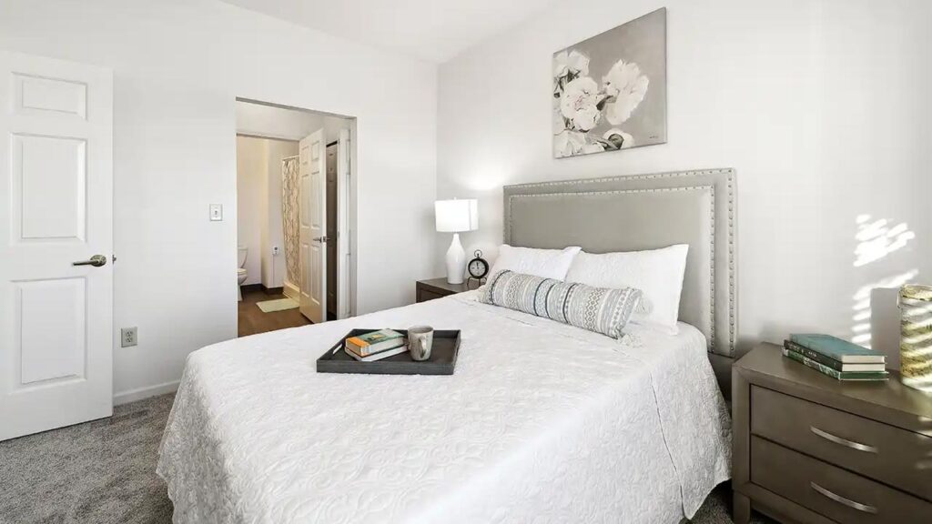 A bright, clean bedroom with a neatly made bed, white bedding, and gray upholstered headboard. A nightstand holds books and a lamp. A tray with a cup and decor sits on the bed. Artwork of flowers hangs on the wall.