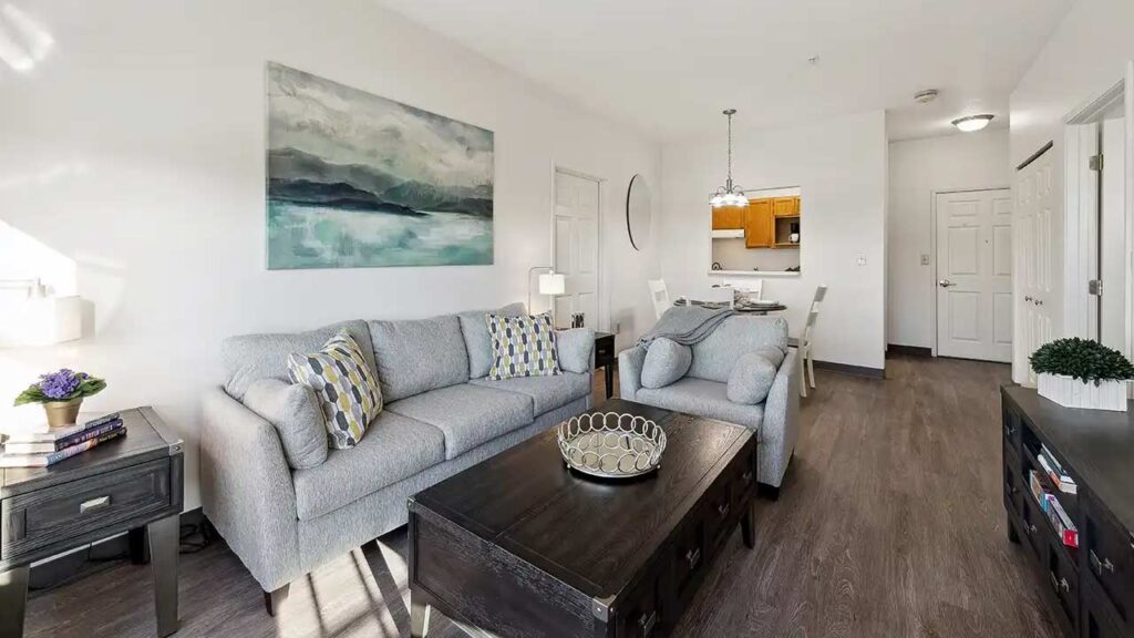 A modern living room with a gray sofa, patterned pillows, coffee table, and side tables, leading to a dining area and kitchen. Artwork and plants decorate the space, which has wood floors and white walls.