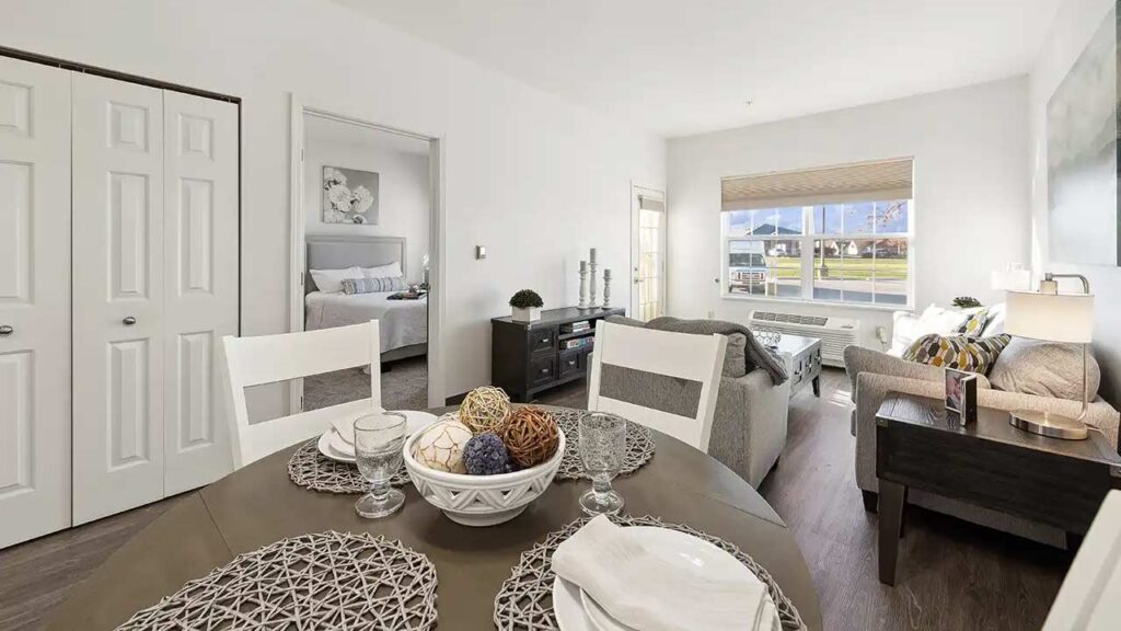 Bright, modern apartment with a dining table in the foreground, a cozy living area with a sofa and TV, and a bedroom visible through an open door. Large window lets in natural light.