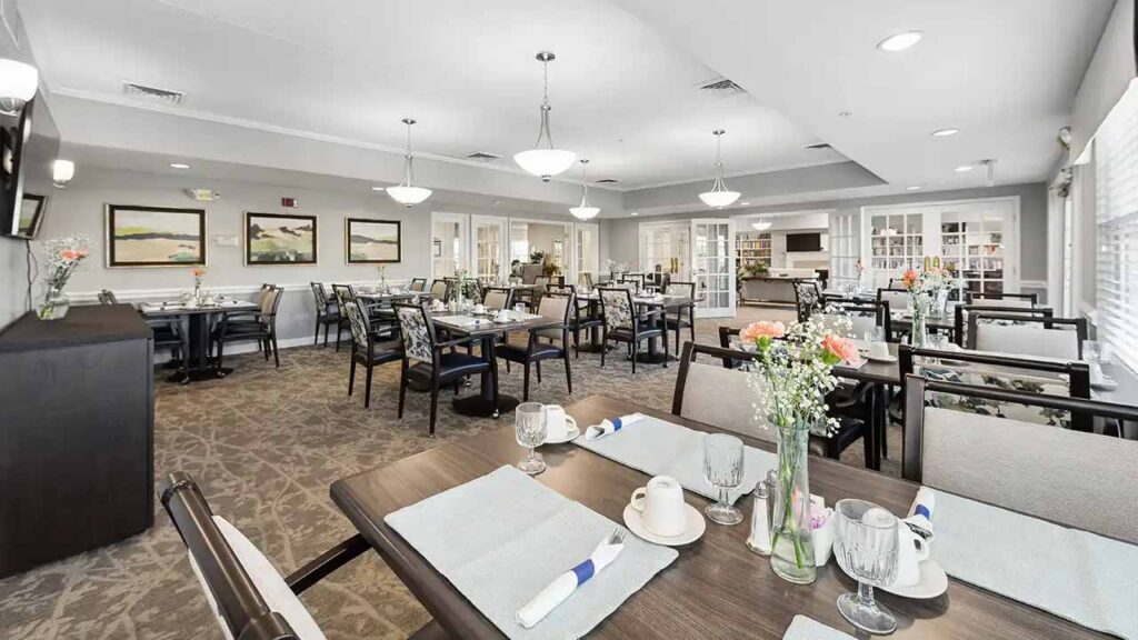 A bright, spacious dining room with neatly set tables, cushioned chairs, and place settings. Decorative flowers, large windows, and framed artwork create a welcoming, airy atmosphere.