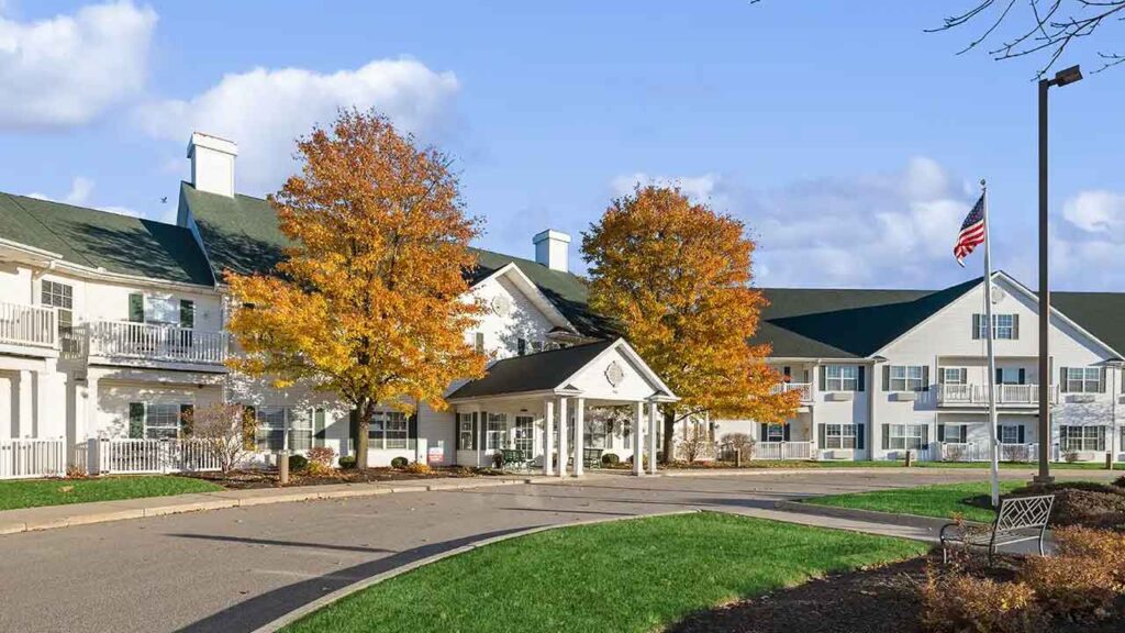 A large, white building with green roofs, a covered entrance, and an American flag out front. Two trees with autumn leaves stand near the driveway under a partly cloudy sky.