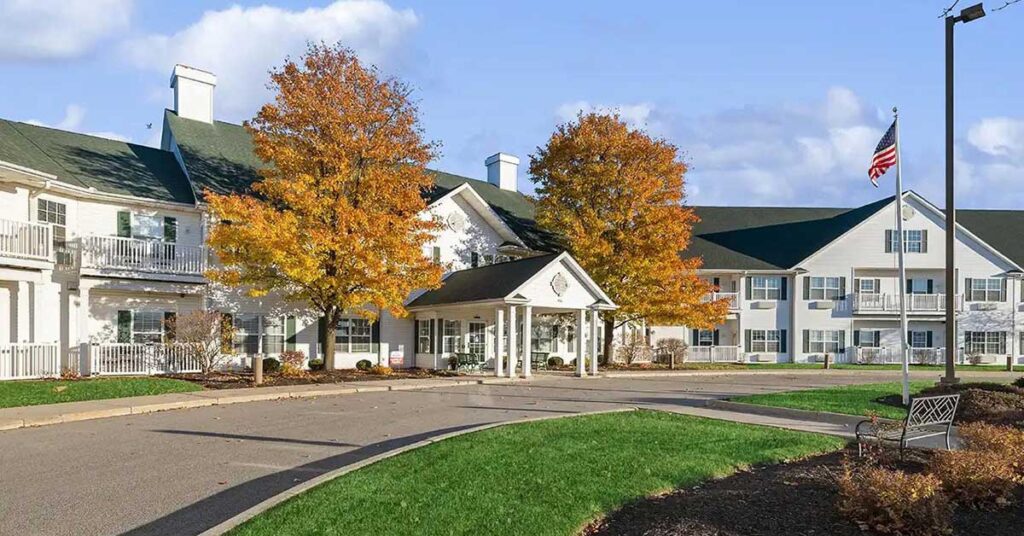 A large white building with green roofs, surrounded by autumn trees with yellow leaves, an American flag on a tall pole, and curved driveways with green grass in front.