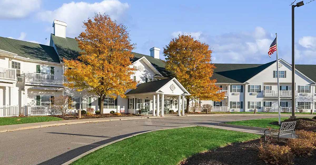 A large white building with green roofs, surrounded by autumn trees with yellow leaves, an American flag on a tall pole, and curved driveways with green grass in front.