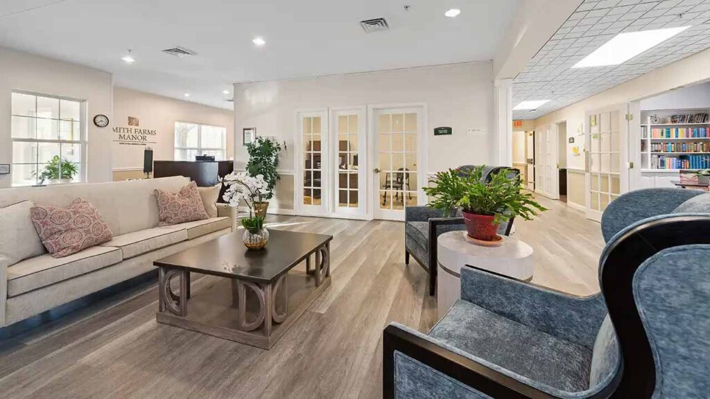 A bright, spacious living room with a beige sofa, armchairs, and a coffee table with flowers, featuring wood floors, potted plants, bookshelves, and glass doors in a retirement or assisted living facility.
