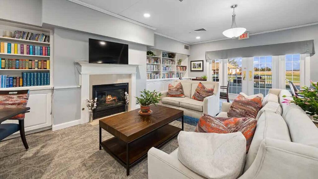 A cozy living room with beige sofas, colorful cushions, a coffee table with a plant, a fireplace, built-in bookshelves, a wall-mounted TV, large windows, and glass doors letting in natural light.