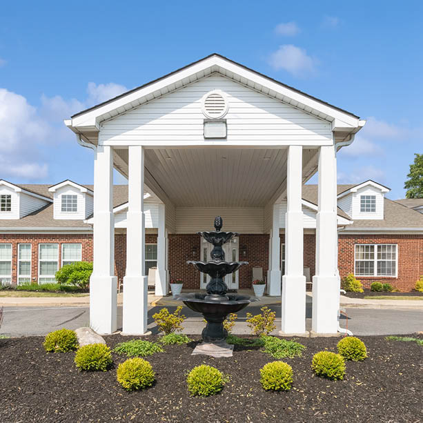 A brick building with white pillars and a triangular roof overhang, featuring a black tiered fountain and neatly trimmed bushes in the landscaped area in front.