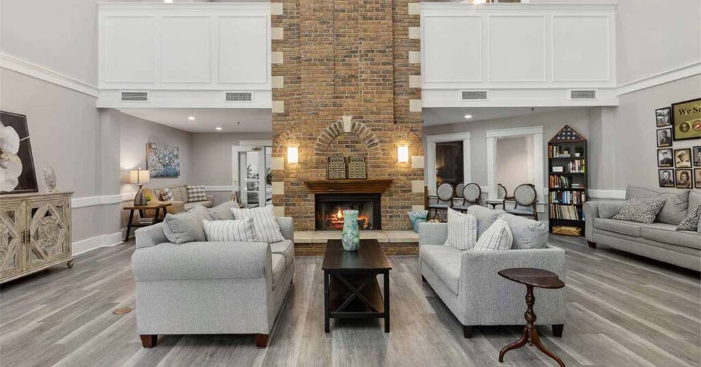 A spacious living room with gray sofas, a wooden coffee table, and a large brick fireplace as the centerpiece. Light wood floors, white walls, and decorative accents create a bright, welcoming atmosphere.