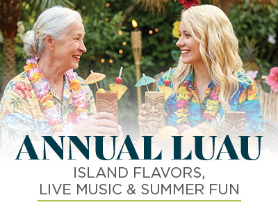 Two women in colorful leis and tropical shirts smile and hold tiki drinks at an outdoor luau, with palm trees and tiki torches in the background. Text reads: