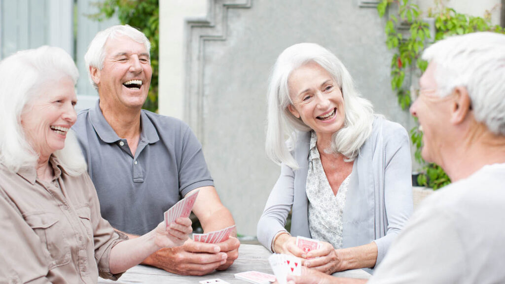 Four older adults with white hair are sitting outdoors around a table, smiling and laughing while playing cards together.