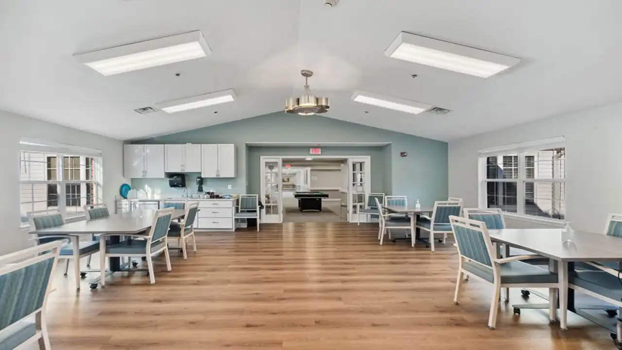 A spacious, well-lit common room with several tables and chairs, wood flooring, a kitchenette at the back, white cabinetry, and large windows allowing natural light to enter. A pool table is visible in an adjacent room.