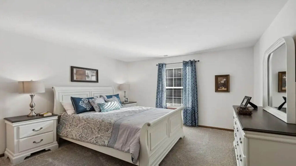 A bright bedroom with a white bed, blue and gray bedding, two nightstands with lamps, a dresser with a mirror, blue patterned curtains on a window, and framed wall art. The room has beige carpet and white walls.