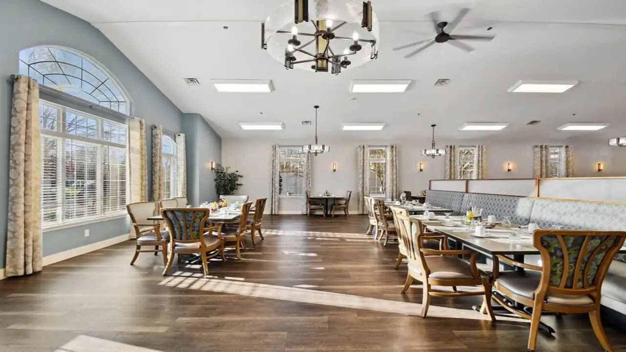 Bright dining area with wooden floors, multiple tables and chairs, large windows with curtains allowing natural light in, and modern chandeliers hanging from the ceiling.