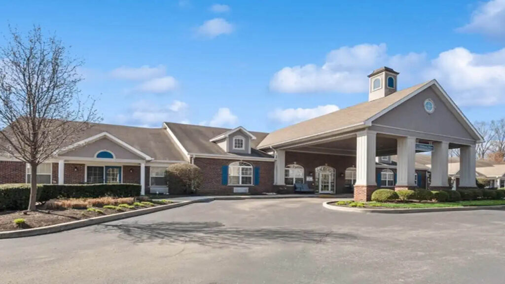 A large, single-story brick building with white columns and a covered driveway, surrounded by neatly trimmed shrubs and trees under a blue sky with scattered clouds.