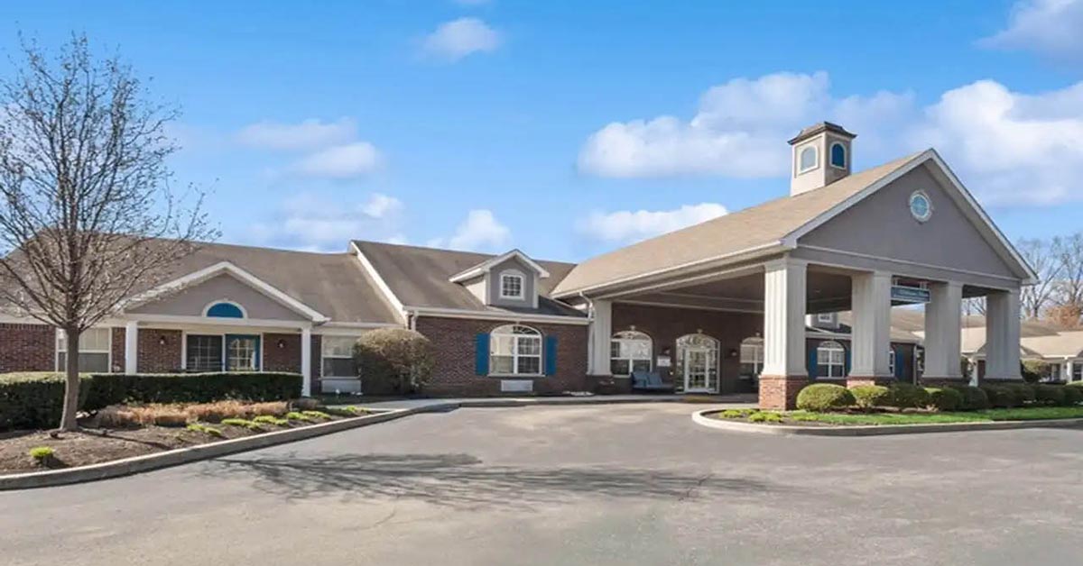 A large, single-story building with brick and beige siding, a covered entrance supported by columns, landscaped shrubs, and a clear blue sky in the background.