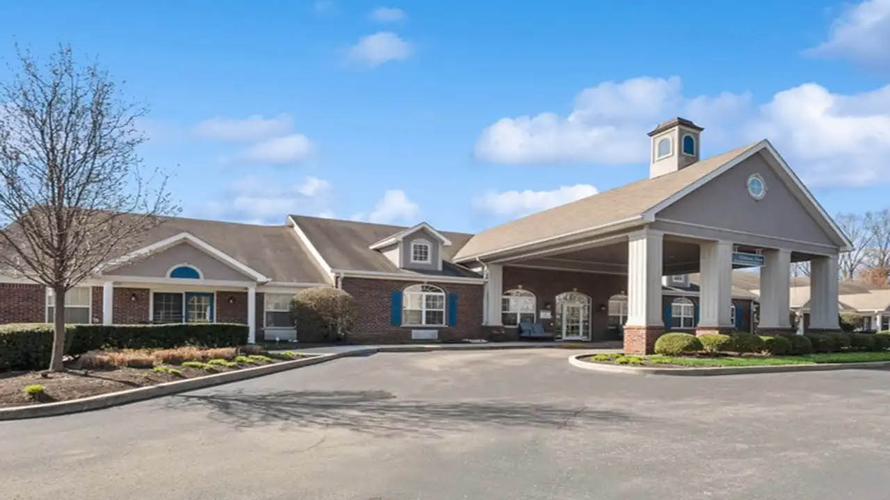 A large, single-story brick building with white columns and a covered driveway, surrounded by neatly trimmed shrubs and trees under a blue sky with scattered clouds.
