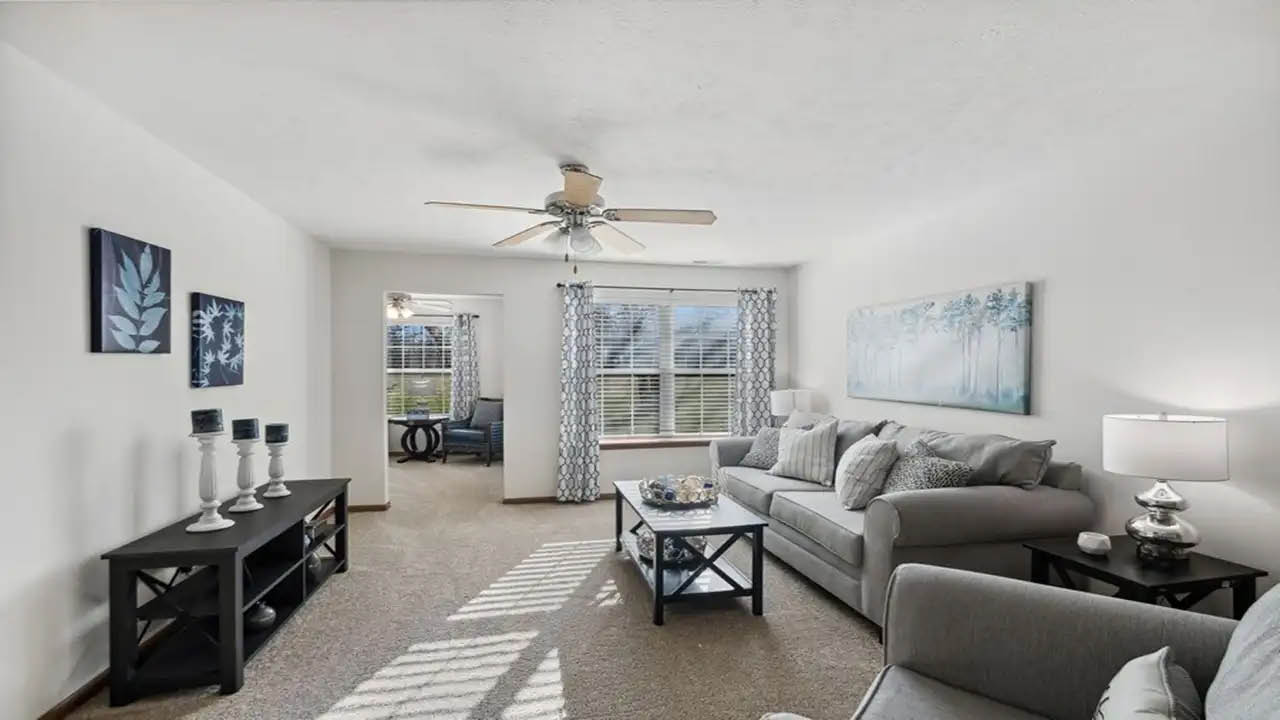 A bright living room with neutral walls, a ceiling fan, gray sofas, a black coffee table, matching end tables with lamps, and wall art. Sunlight streams in through large windows, creating a cozy atmosphere.