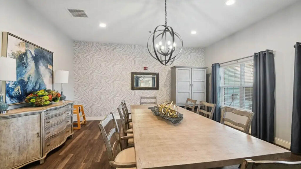A dining room with a long wooden table, eight chairs, a modern chandelier, a sideboard with lamps and flowers, a large abstract painting, and a window with gray curtains letting in natural light.