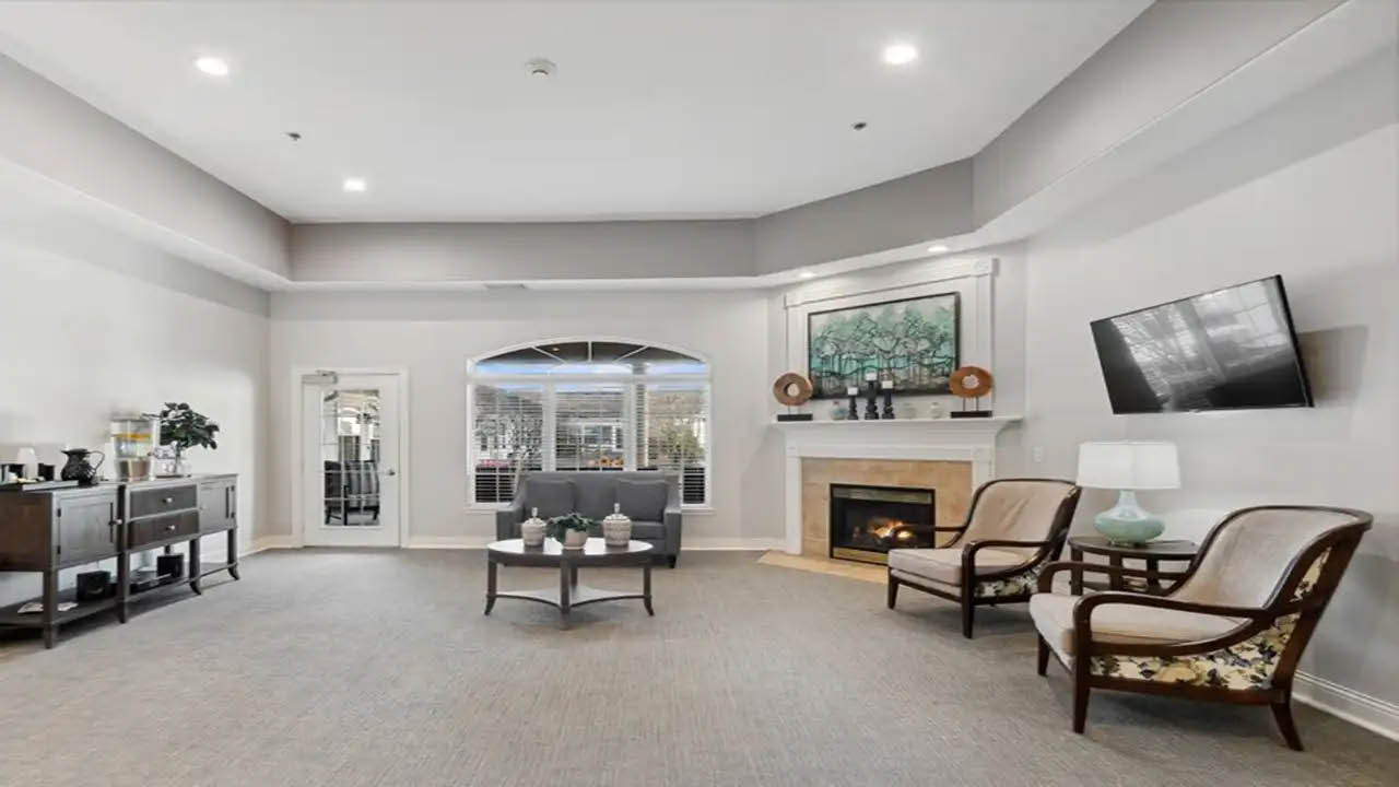 A bright living room with neutral walls, two armchairs, a fireplace with art above it, a wall-mounted TV, and a console table with decor. Large windows and a glass door let in natural light.