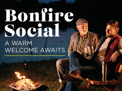 Two men sit by a small bonfire near a lake at dusk, smiling and holding mugs. Text reads: “Bonfire Social. A warm welcome awaits.”.