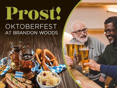 Two men clinking beer glasses and smiling. Nearby, plates of sausages, pretzels, and sauerkraut. Text reads: “Prost! Oktoberfest at Brandon Woods.”.