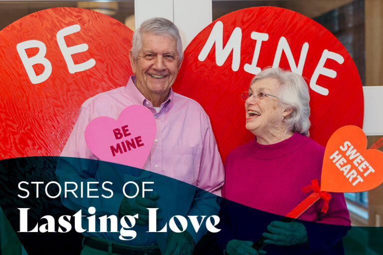 An older man and woman smile together, holding heart-shaped signs reading "BE MINE" and "SWEETHEART," in front of large red hearts that say "BE MINE." Text overlay reads “Stories of Lasting Love.”.