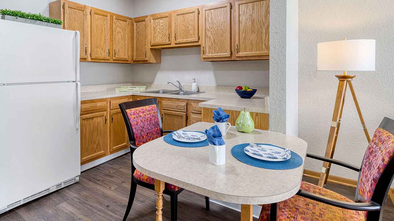 Kitchen and dining area featuring wooden cabinets, a white refrigerator, and a table set for two with blue placemats and patterned chairs.