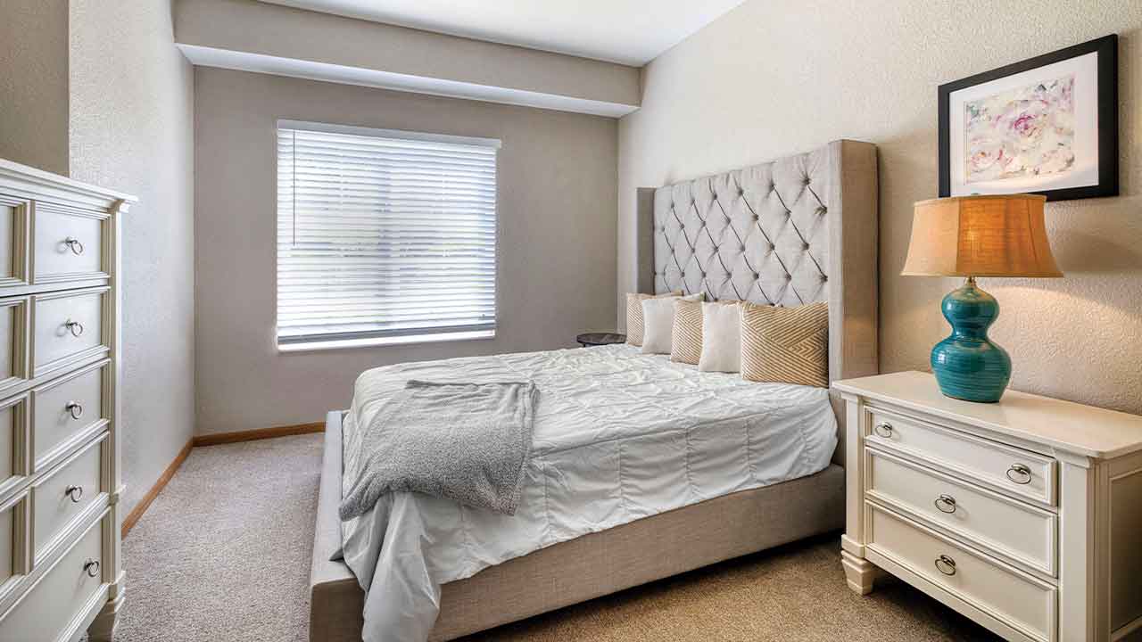 Bedroom with a tufted headboard bed, two white wooden dressers, a bedside lamp with a blue base, and a framed floral print on the wall.