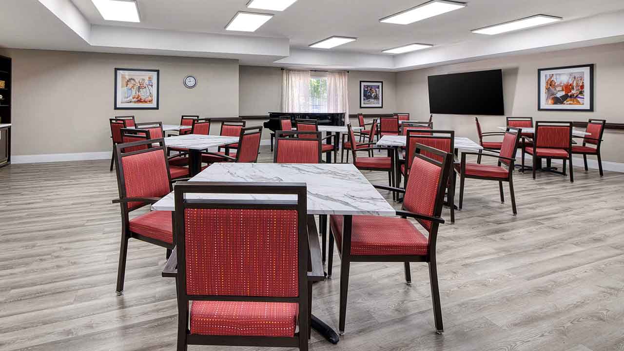 Dining area with multiple square tables and red upholstered chairs arranged on a light wood floor, with framed artwork on the walls and a piano in the background.