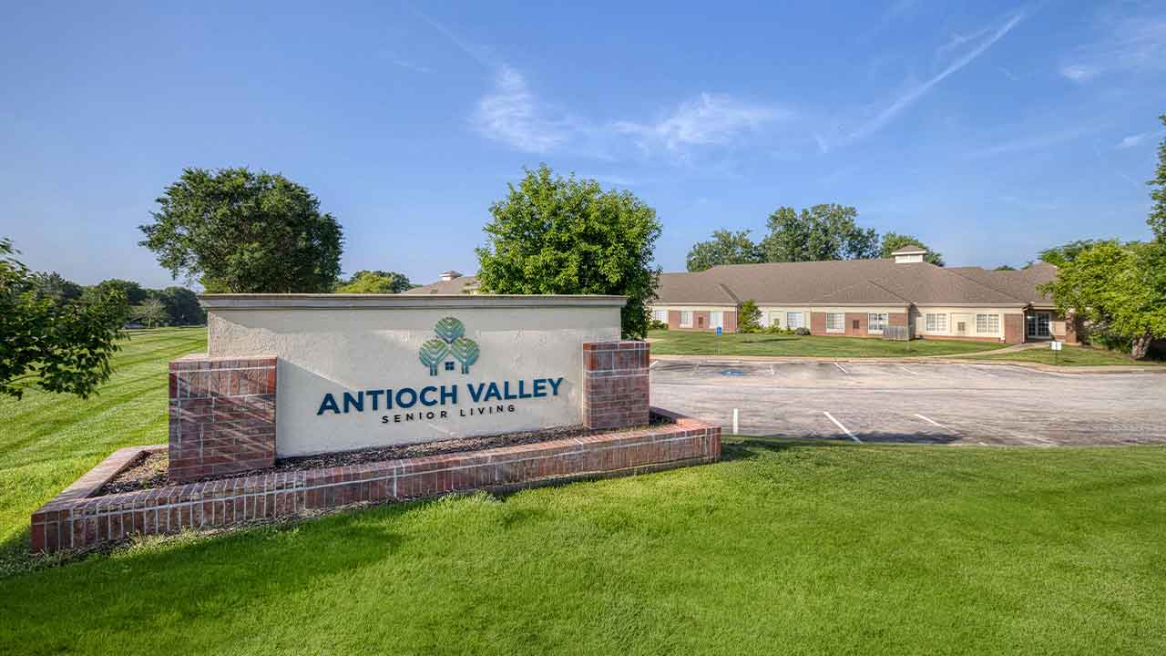 A brick and stone sign for Antioch Valley Senior Living in front of a parking lot and a low-rise residential building.