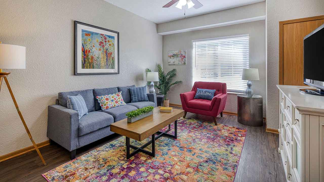 Living room with a gray sofa, a red armchair with a “Welcome” pillow, a wooden coffee table, and a colorful patterned area rug.