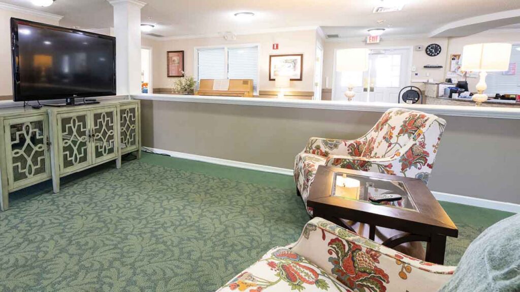 A cozy living room with patterned armchairs, a wooden coffee table, and a large flat-screen TV on a decorative cabinet. The room has green carpet, soft lighting, and neutral walls with artwork and lamps.