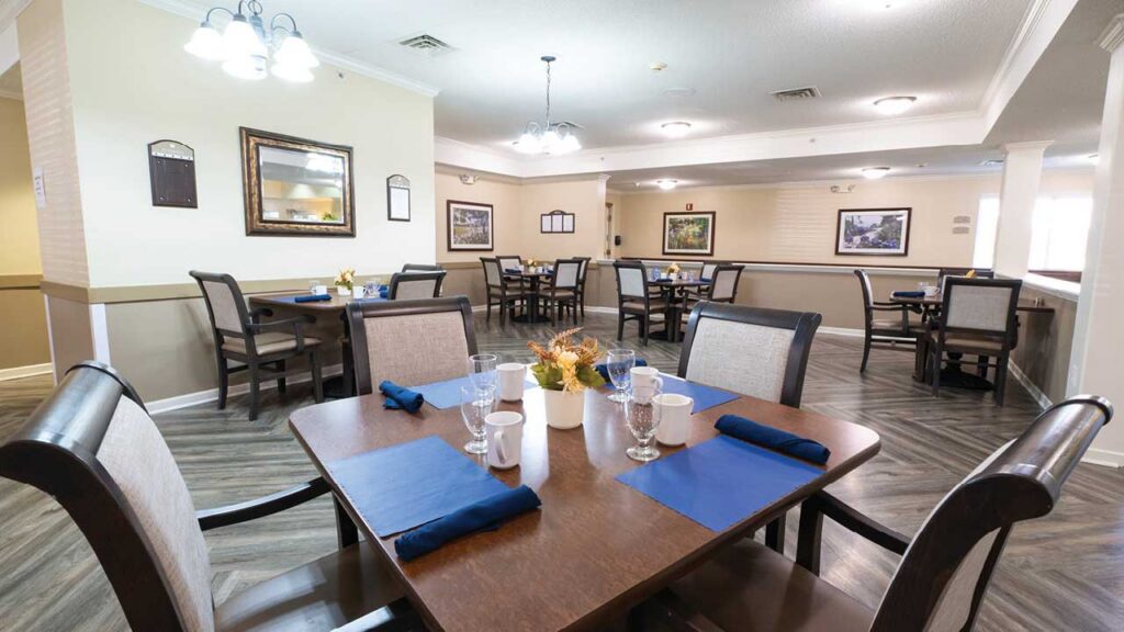 A bright, spacious dining area with several wooden tables and chairs, set with blue napkins, glasses, and cutlery. Framed artwork decorates the walls, and ceiling lights provide a warm atmosphere.