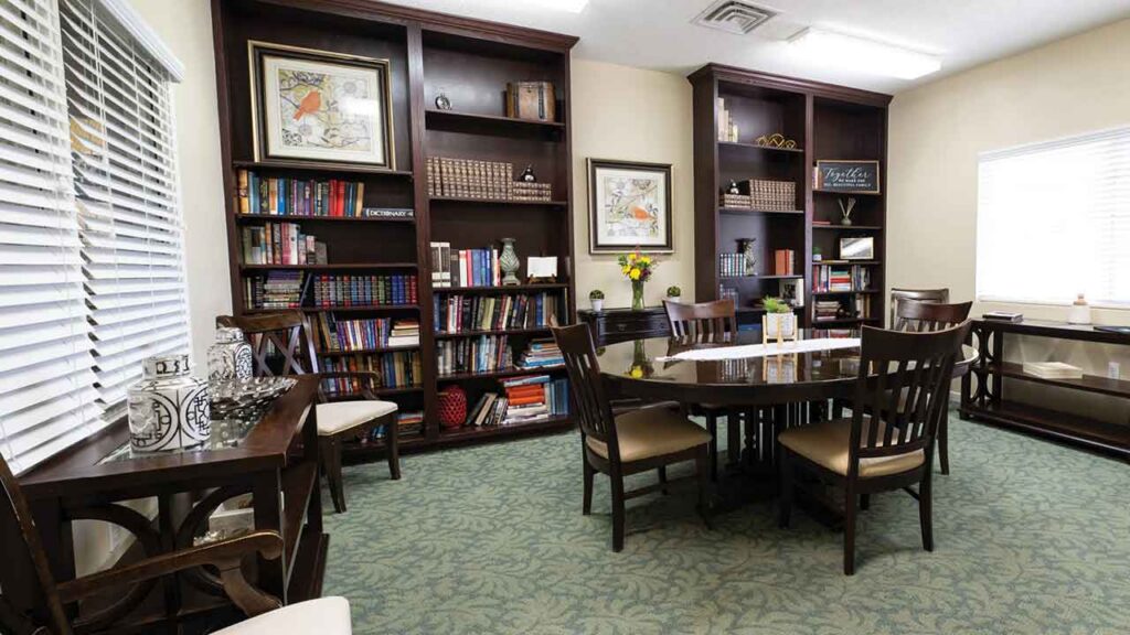 A library room with dark wooden bookshelves, a large table with chairs, books, framed art, flowers, and windows with blinds letting in natural light. The room has a green patterned carpet.