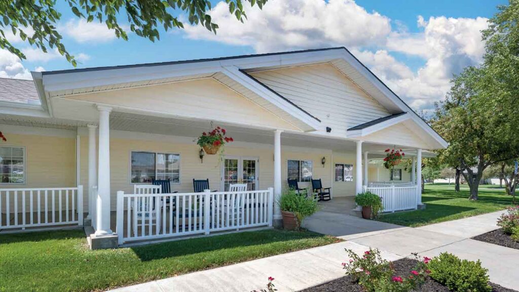A single-story building with a white porch, hanging flower baskets, rocking chairs, and green lawns, set under a partly cloudy blue sky.