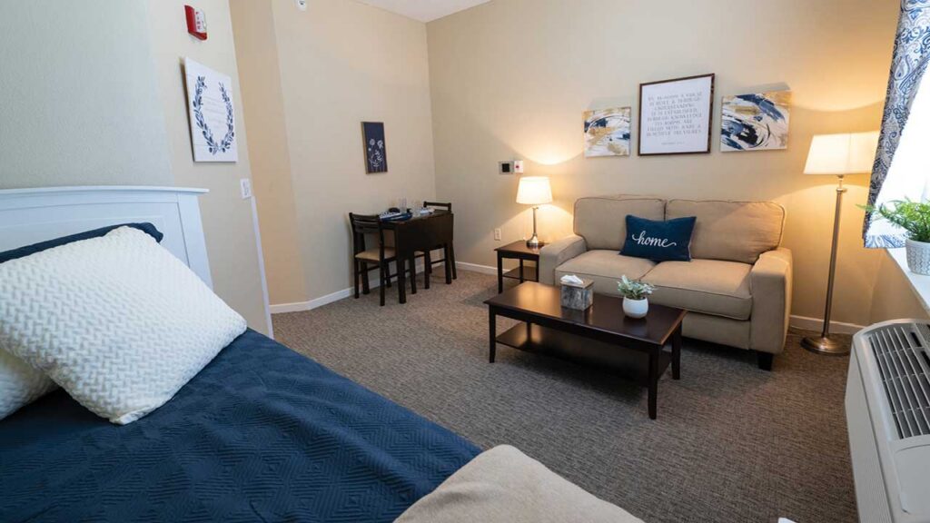 A cozy, well-lit studio room with a neatly made bed, a small sofa and coffee table, a desk with two chairs, wall art, a lamp, and a window with patterned curtains. The space is decorated in neutral tones.