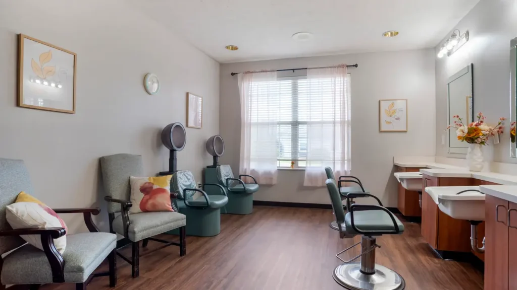 A bright, tidy salon room with three styling chairs, two hooded hair dryers, cushioned waiting chairs, a large window with sheer curtains, and wall art. The space has wood flooring and a countertop with a vase of flowers.