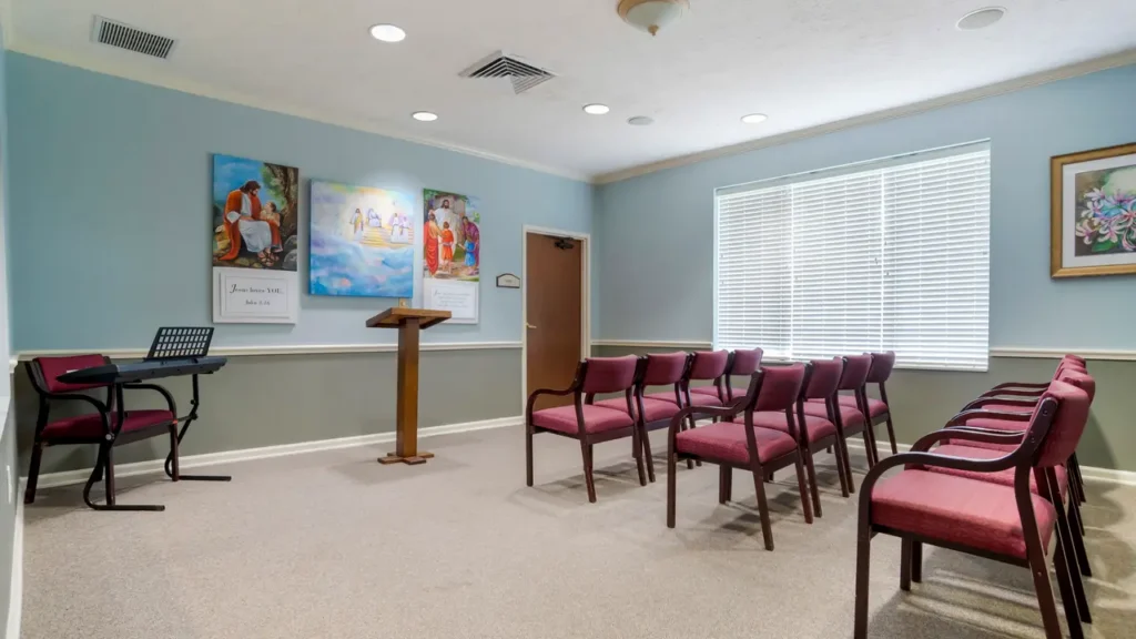 Small chapel room with rows of red cushioned chairs facing a wooden lectern. Religious paintings and a sign hang on the blue wall. Sunlight streams through a window with blinds on the right.