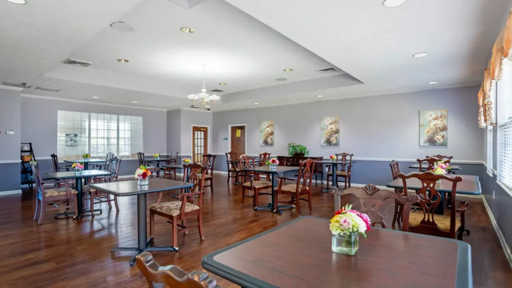 A bright, spacious dining room with wooden floors, several tables and chairs, floral centerpieces, wall art, large windows, and soft lavender walls. Natural light fills the room, creating a welcoming atmosphere.