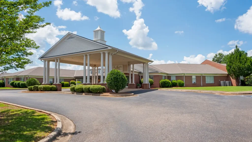 A single-story brick building with a large covered entrance supported by white columns, surrounded by neatly trimmed bushes and trees under a blue sky with scattered clouds.